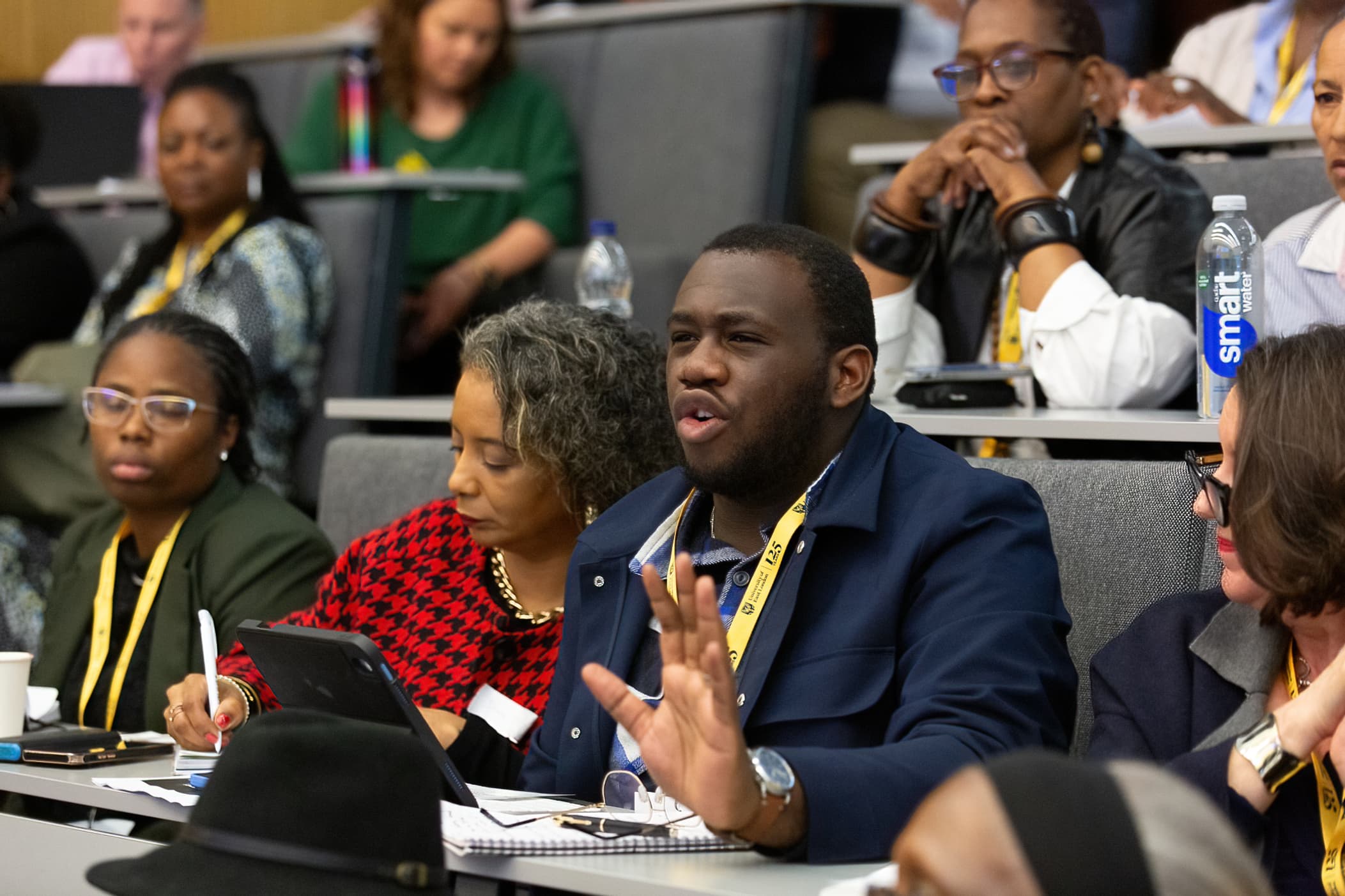 A delegate gesturing as he raises a point from the floor of a lecture theatre at an APA conference, with others listening around him