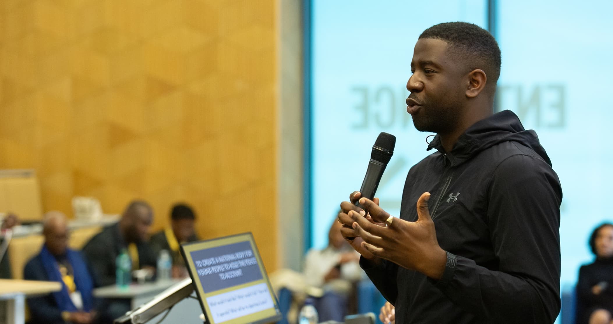 A young man speaking into a microphone at an APA conference, beside a screen reading "To create a national body for young people to hold the police to account"