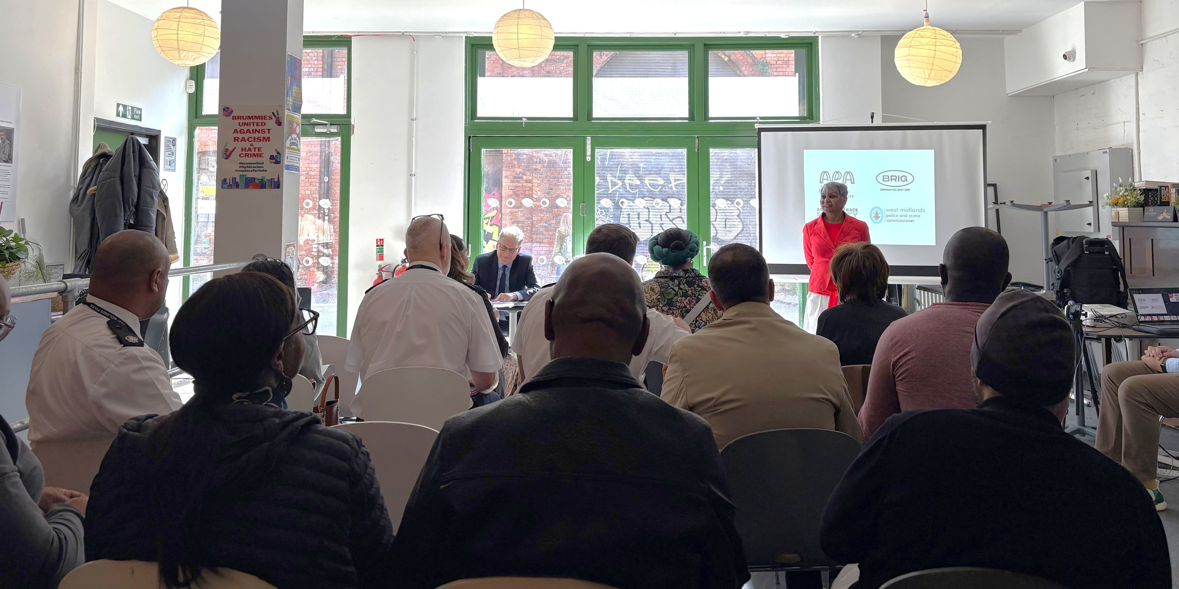 A speaker presenting to a seated audience that includes police officers, beside a screen showing the APA, Birmingham Race Impact Group, Action for Race Equality and West Midlands Police and Crime Commissioner logos